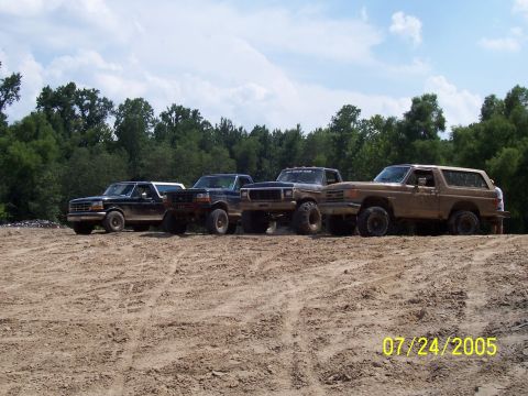 1987 Ford Bronco - A lineup of four Ford vehicles, including a tan Bronco and three trucks, are parked on a dirt track under a cloudy sky.