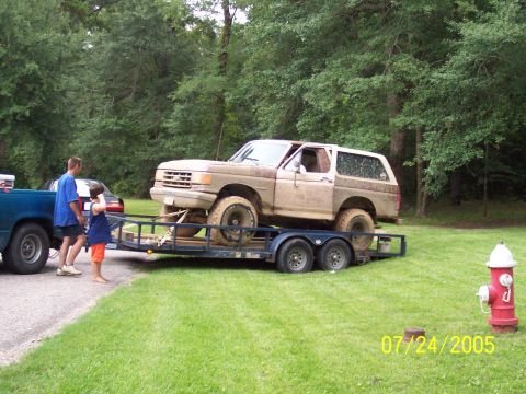 1987 Ford Bronco - Mud-caked full-size Ford Bronco strapped onto a blue tandem-axle open trailer behind a teal pickup, with a man and a child standing alongsi…