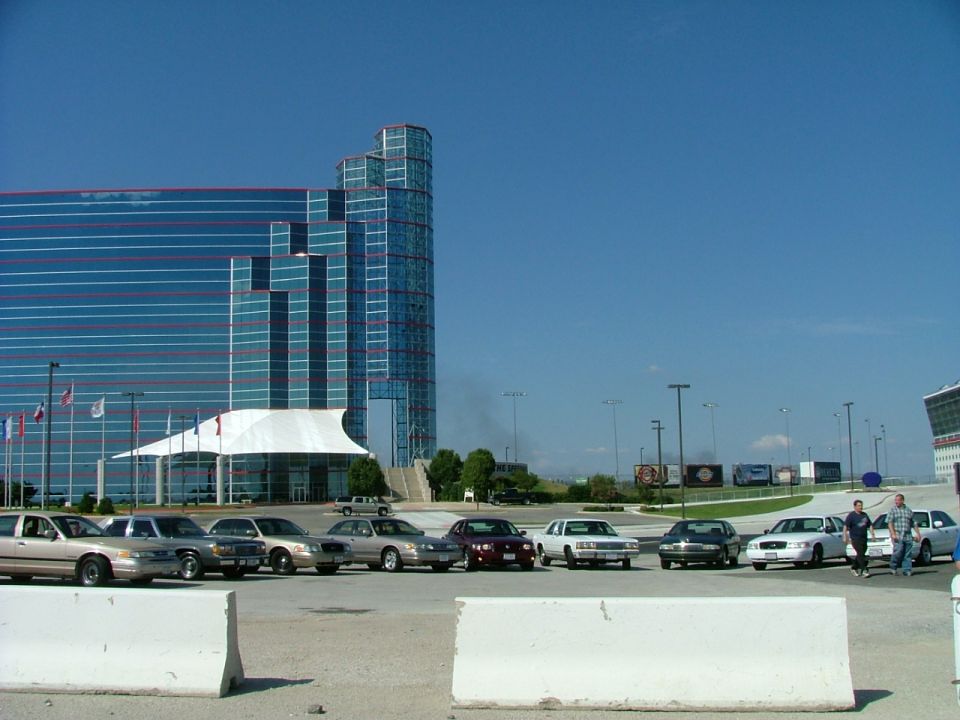 Several tan and silver Ford Crown Victorias parked in a lot with a modern building in the background