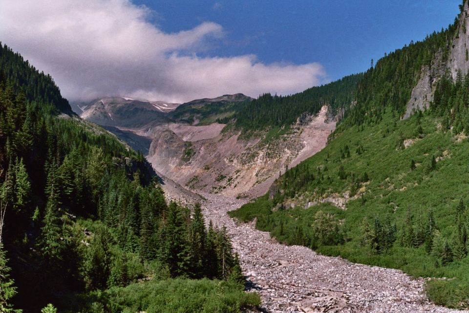 Mountain valley with pine trees and a rocky riverbed, under a partly cloudy sky. Supermotors.net logo visible