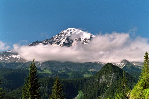 1979 Ford Bronco - A mountain landscape with a snow-covered summit partially obscured by clouds, set against a backdrop of dense forests and rolling hills.