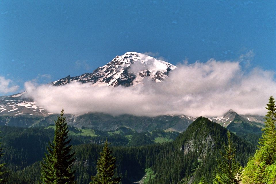 Snow-capped mountain peak shrouded in clouds, surrounded by evergreen forests and green hills under a blue sky