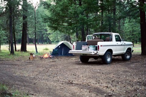 1979 Ford Bronco - Campsite scene with a white Ford Bronco, tent, and campfire in a forest setting.