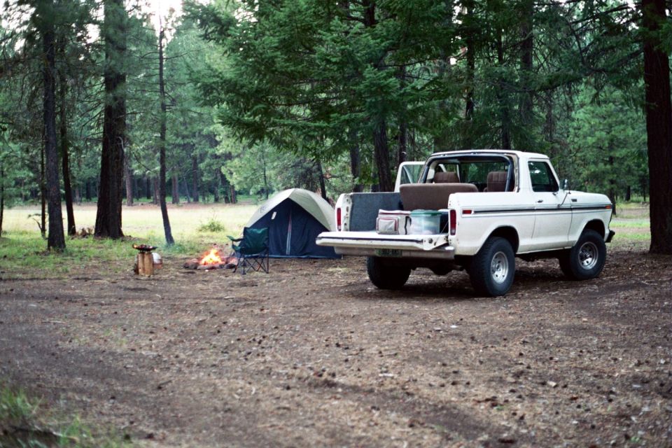 White 1978 Ford Bronco with tailgate down, parked near a tent and campfire in a wooded area