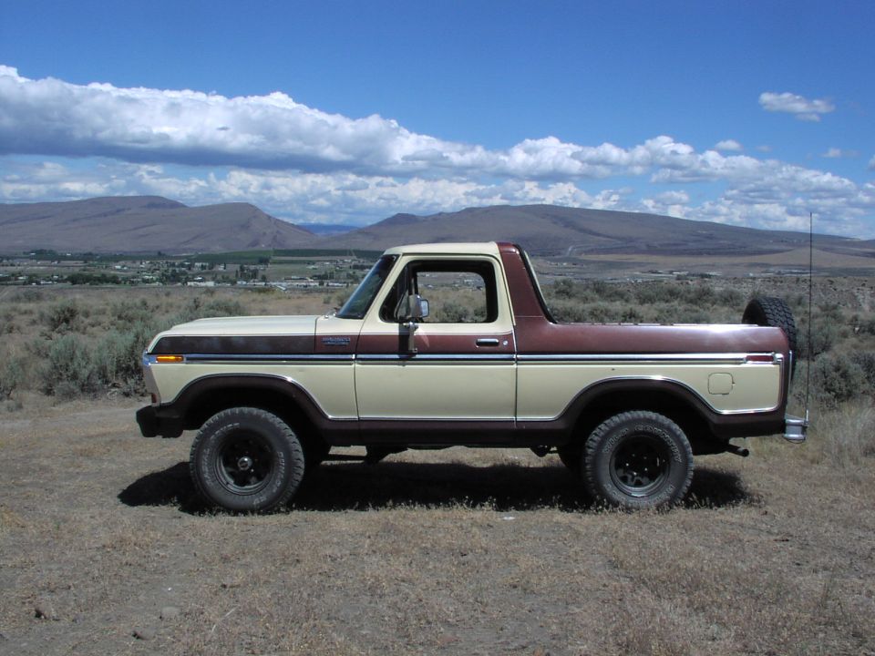 Side view of a tan and brown Ford Bronco parked on dry grass with hills and clouds in the background