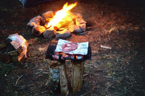 1979 Ford Bronco - A steak and onions are grilled over a campfire, with a stack of firewood nearby.