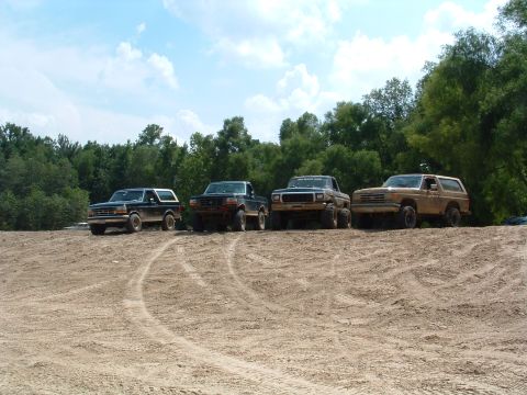 1993 Ford Bronco - Four Ford Bronco SUVs and trucks are parked on a dirt hill under a cloudy sky.