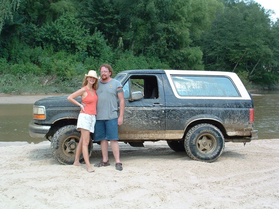 1993 Ford Bronco parked on sandy riverbank with two people standing next to it