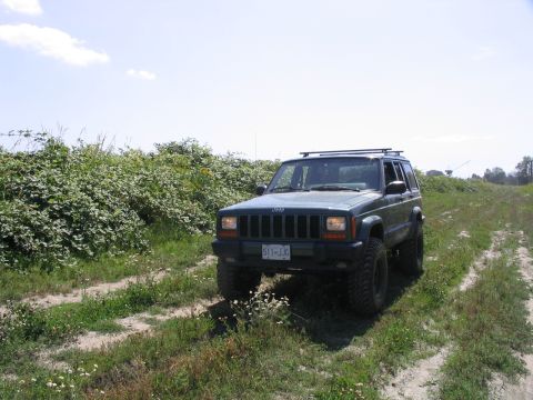 2000 Jeep Cherokee - A dark green Jeep Cherokee drives on a dirt path surrounded by vegetation under a partly cloudy sky.