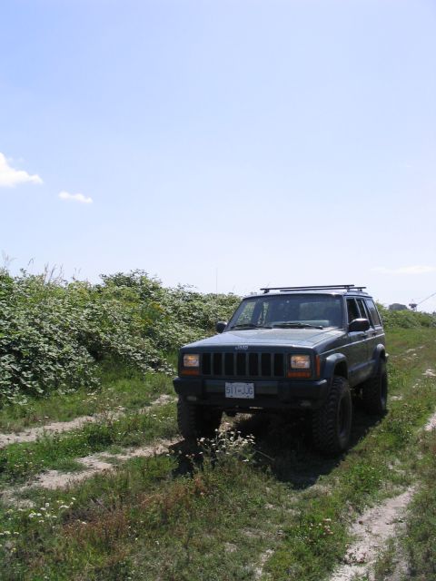 2000 Jeep Cherokee - Jeep Cherokee parked on a dirt road with green foliage under a blue sky.