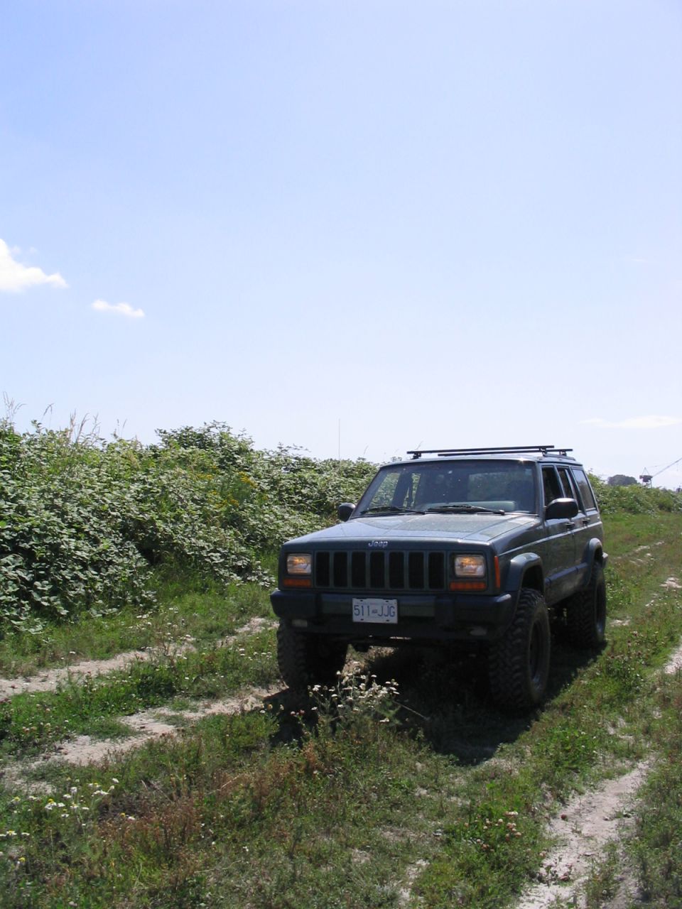 Dark green Jeep Cherokee parked on a dirt road with overgrown bushes on the left. License plate reads 511-JJG