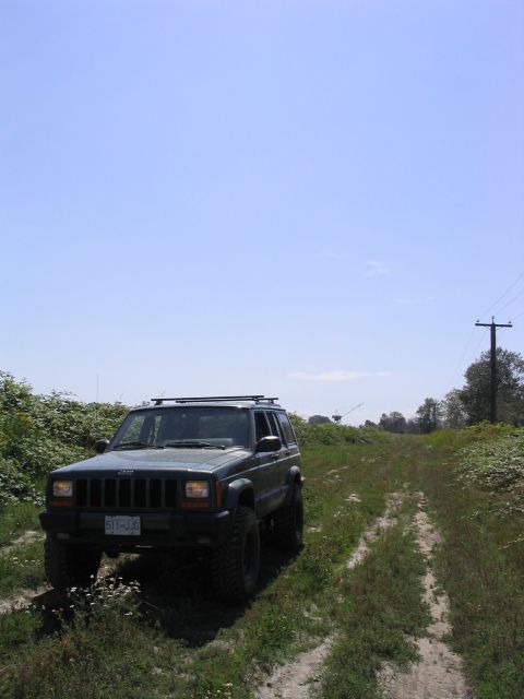 2000 Jeep Cherokee - A Jeep Cherokee XJ is parked on a dirt path surrounded by greenery under a clear blue sky.