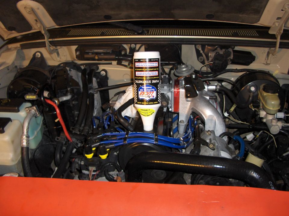 Engine bay with upside-down Lucas Synthetic Oil Stabilizer bottle draining into oil fill; blue plug wires visible.