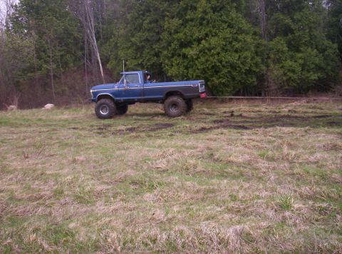 1984 Ford Bronco - A blue lifted pickup truck is being towed across a grassy field with trees in the background.