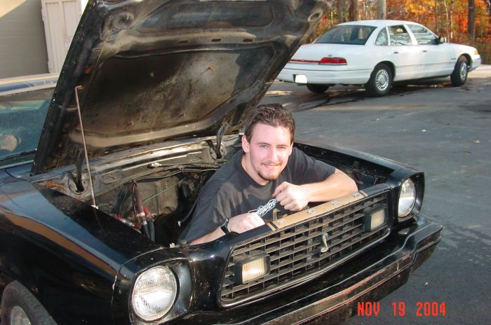Man leaning over grille of black Mustang II with hood up, white sedan behind.