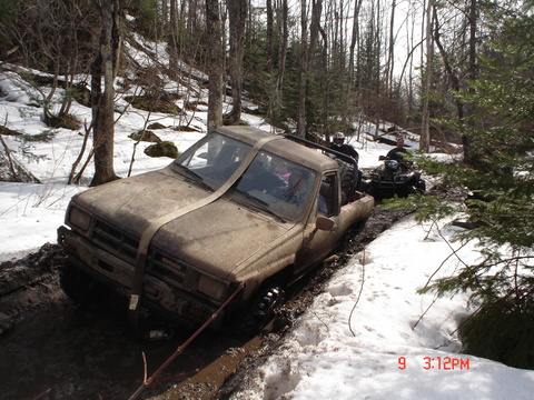 1995 Ford Bronco - A pickup truck is stuck in a muddy, snowy trail while an ATV follows behind in a wooded area.