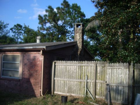 1978 Ford Bronco - A residential scene a brick house with a prominent stone chimney and a weathered wooden fence under a blue sky.