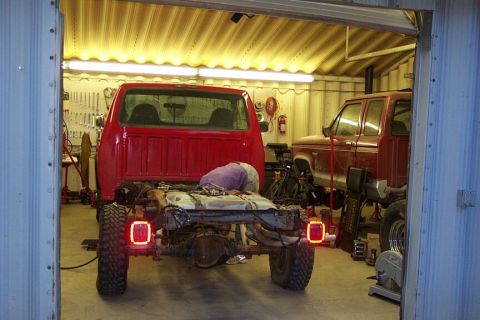 1990 Ford Bronco - A red truck chassis is being worked on in a garage, with another red truck visible in the background.