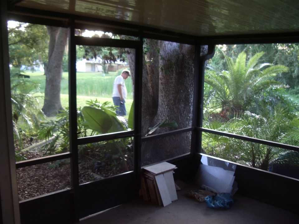 screened porch looking out at a man in a white shirt and blue shorts walking in a grassy yard with trees and palm trees