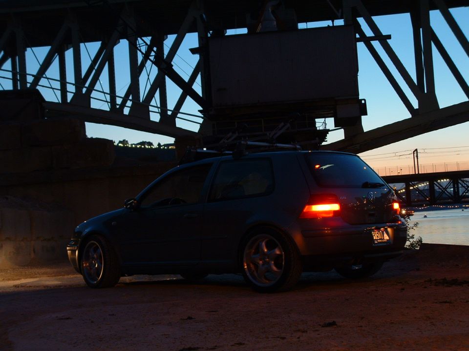 Dark hatchback parked under a bridge at dusk, rear view. Has a roof rack