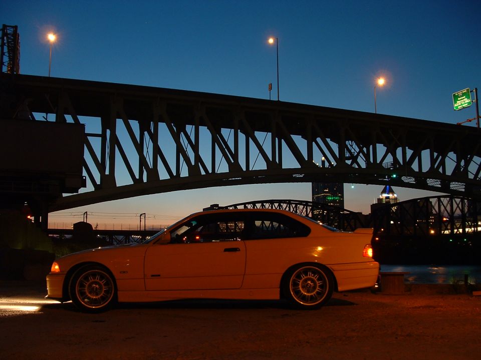 Yellow coupe parked under a bridge at dusk, side view