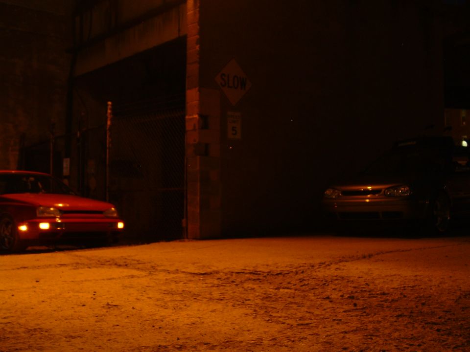 Red car on left, dark car on right, in a dark alley with a SLOW sign