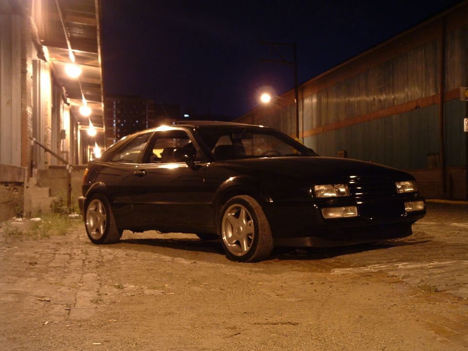 Black coupe parked on gravel at night, with building lights in background