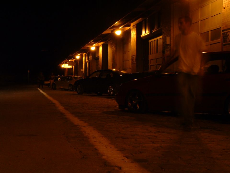 Several cars parked in a row at night next to a building with lights. A person is walking near the front car