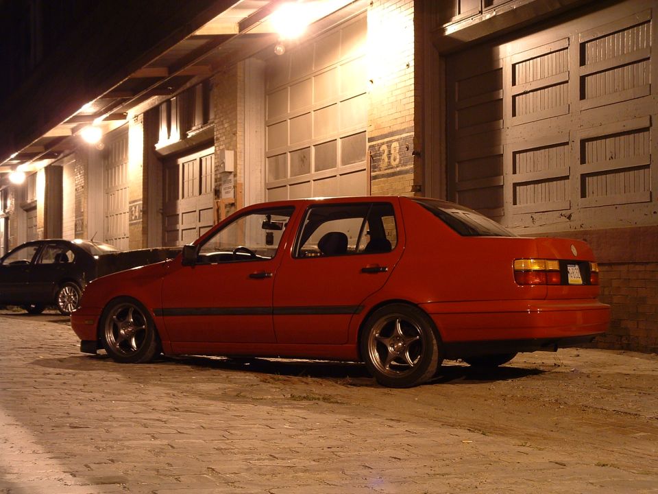 Red sedan parked on cobblestone street at night, side view. Another black car is visible behind it