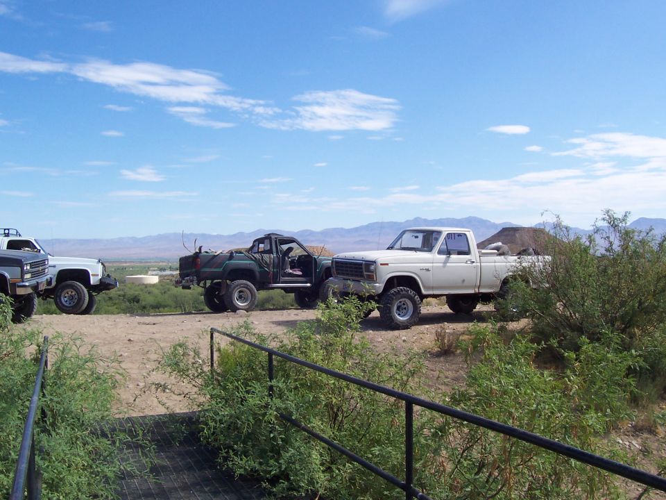 Lifted square-body Chevy, green flatbed truck and white Ford pickup parked on desert ridge