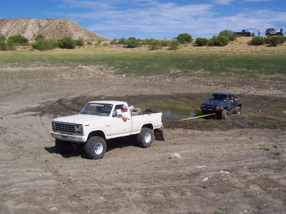 White lifted Ford 4x4 pulling stuck dark truck from mud with yellow strap, mountains behind