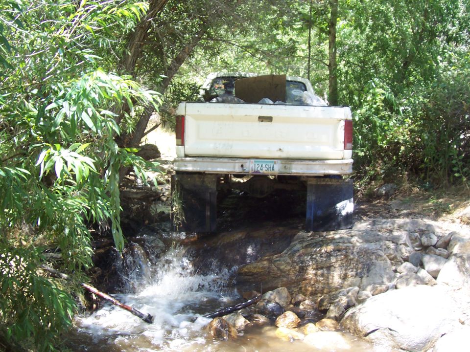 Rear of white Ford pickup fording a shallow rocky creek under overhanging trees