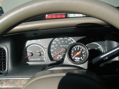 2000 Ford Crown Victoria - Instrument cluster viewed through a tan steering wheel, showing an aftermarket black-faced 8,000-RPM Auto Meter tachometer mounted on a pod…