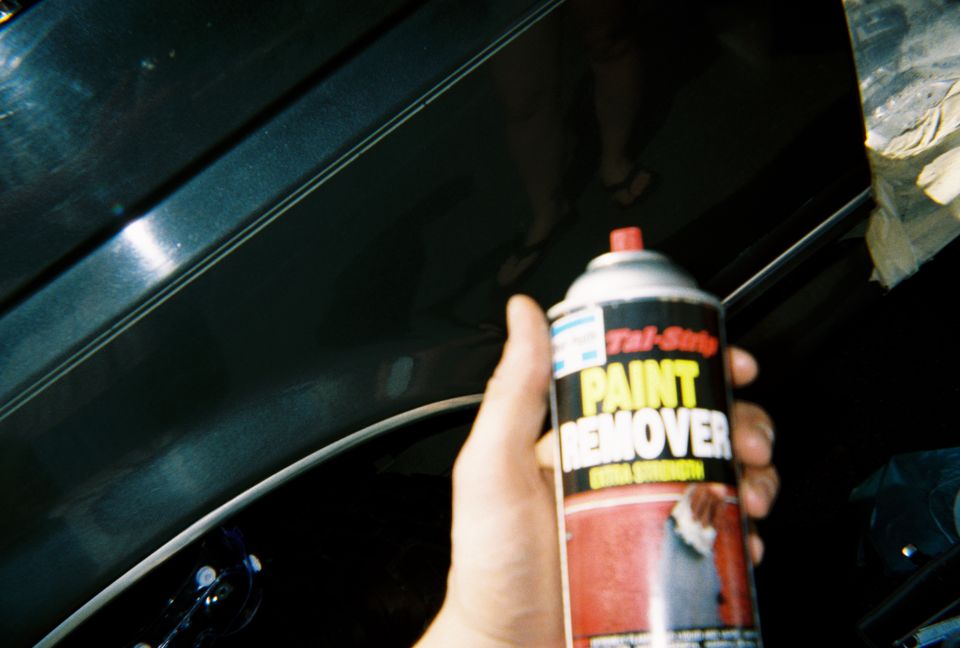 Hand holding a can of Paint Remover spray in front of a dark masked car body panel
