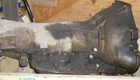 1988 Ford Bronco - A grimy automatic transmission sitting on a wooden pallet or crate in front of a plywood wall, photographed from the side with the bellhous…