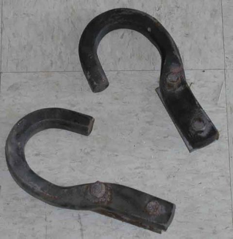 1988 Ford Bronco - A matched pair of heavy forged steel frame-mount tow hooks lying on an OSB plywood floor, painted black with surface rust bleeding through…