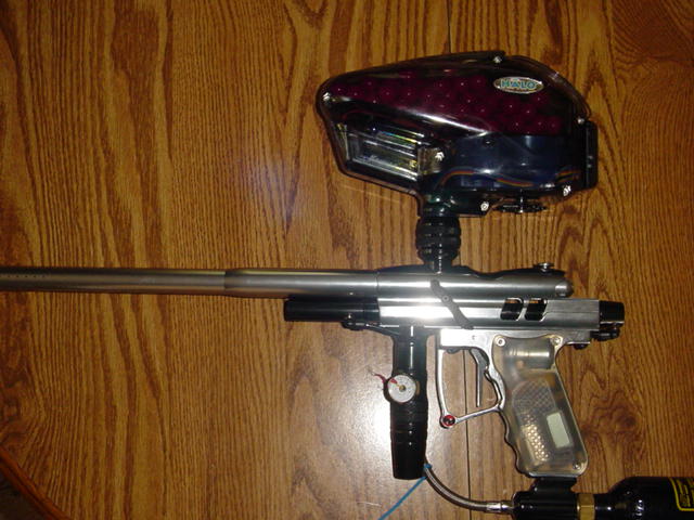 Paintball marker with a clear grip and a red-speckled hopper, resting on a wooden surface