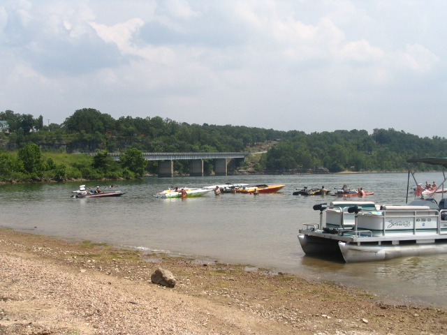 Pontoon boat beached on gravel shore with colorful boats rafted up near a bridge