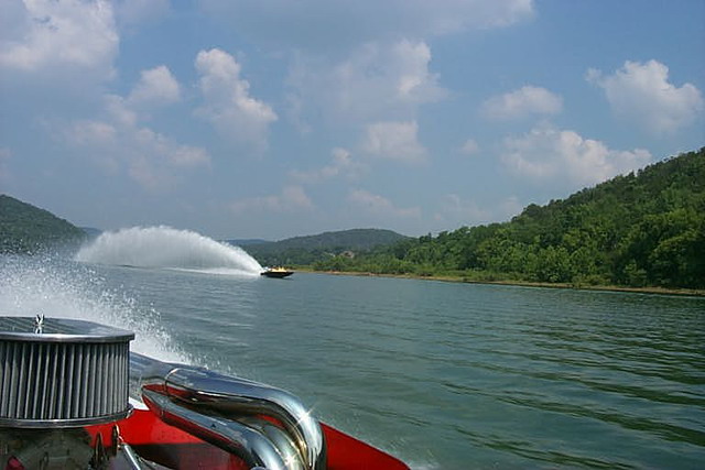 Boat engine and exhaust pipes in foreground, another boat creating a large wake in the distance on a lake