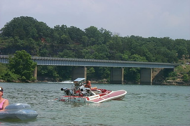 Red and white flat-deck speedboat with blown exposed engine idling near a lake bridge
