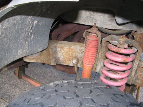 1978 Ford Bronco - The underside of a vehicle shows a rusty suspension system with red shocks and springs, along with exhaust headers.