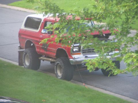 1978 Ford Bronco - A lifted red 1978 Ford Bronco with a white top is seen from the front-right angle, partially obscured by foliage.