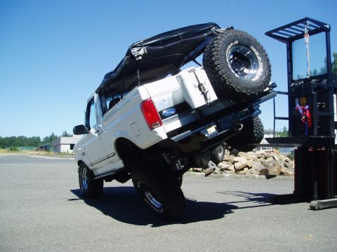 1995 Ford Bronco - A white Ford Bronco is being lifted by a forklift, showing off its suspension and off-road modifications.