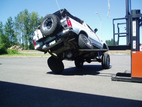 1995 Ford Bronco - A white SUV is precariously lifted by a forklift, revealing its modified undercarriage and oversized tires.