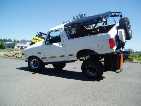 1995 Ford Bronco - White 1995 Ford Bronco is lifted on orange blocks, with a yellow Jeep visible behind it.