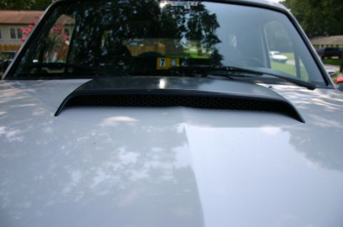 1994 Ford Bronco - Close-up across a silver hood at a black cowl-mounted hood scoop with a honeycomb mesh insert, photographed from the front of the vehicle l…