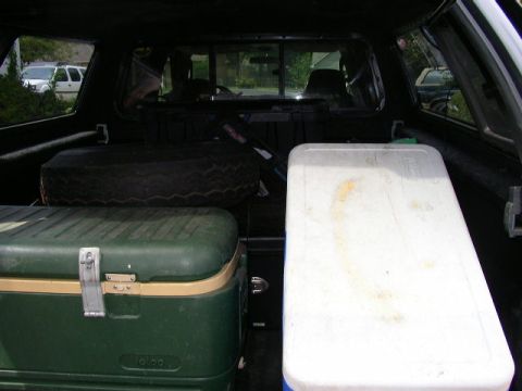 1995 Ford Bronco - The interior of a truck bed is shown, containing two coolers and a spare tire, with a white SUV visible through the rear window.