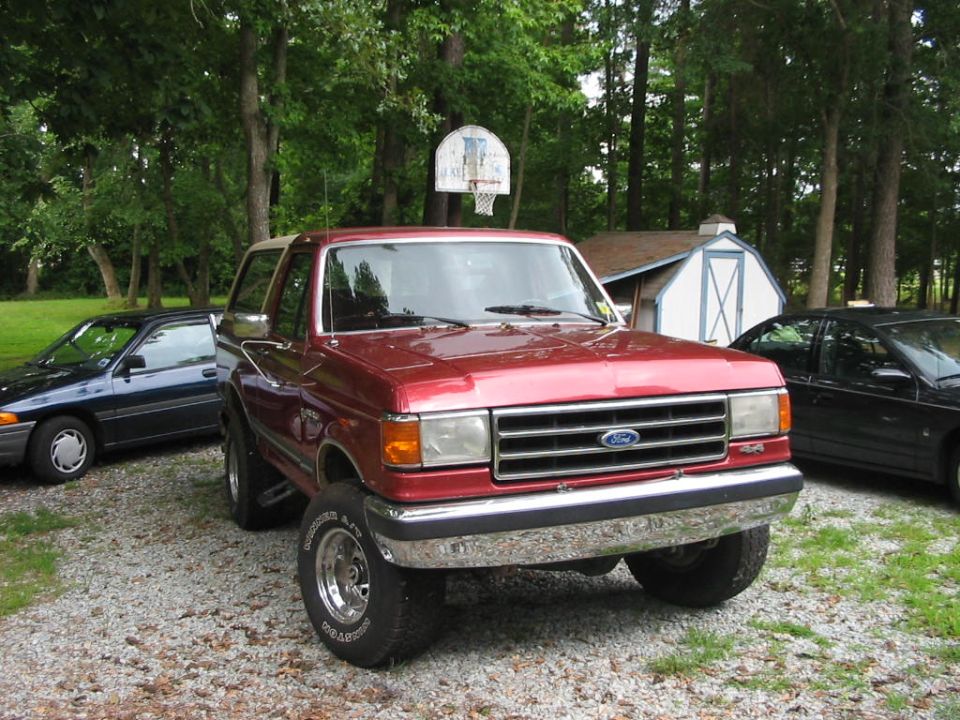 Red Ford Bronco parked on gravel, with a blue sedan and black car behind it. Basketball hoop in background