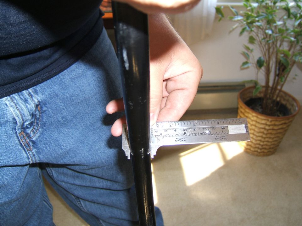 Person in blue jeans measuring a black rod with a caliper, with a potted plant in the background