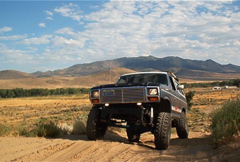 1986 Ford Bronco - This lifted SUV navigates a dusty trail with a mountainous landscape under a partly cloudy sky.